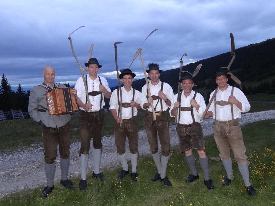 Fünf Plattler mit Sensen und ein Harmonikaspieler auf einer Wiese in der Abenddämmerung